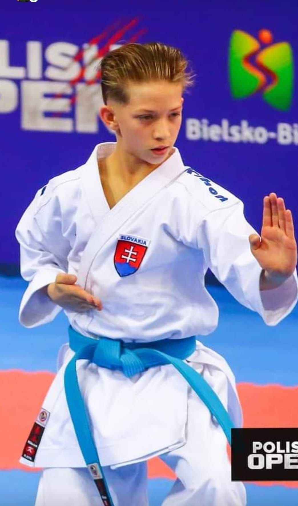 Young karate athlete in white uniform and blue belt performing a kata move at a Polish Open event.